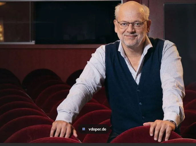 männliche Person mittleres Alter mit Brille in entschlossener Haltung mit beiden Händen gestützt auf samtroter Theaterbestuhlung